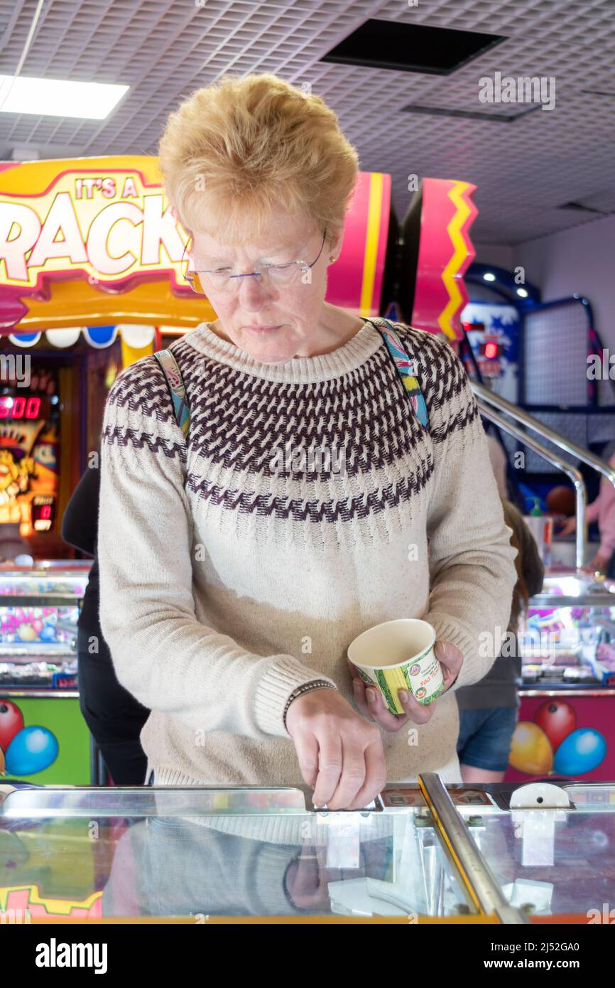 Una mujer de mediana edad alimenta monedas en una máquina de coser de monedas en una sala de juegos electrónicos de entretenimiento del Reino Unido. La mujer está sosteniendo una bañera de plástico llena de cambio para jugar Foto de stock