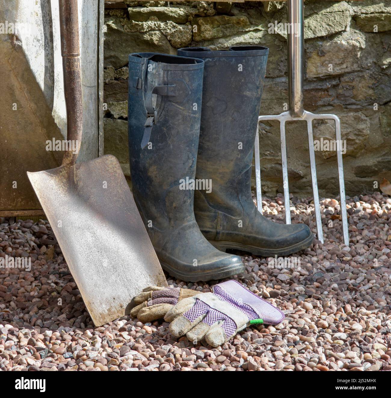 Artículos de jardinería, incluyendo pala grande, tenedor grande, guantes y botas de agua contra la pared de piedra Foto de stock