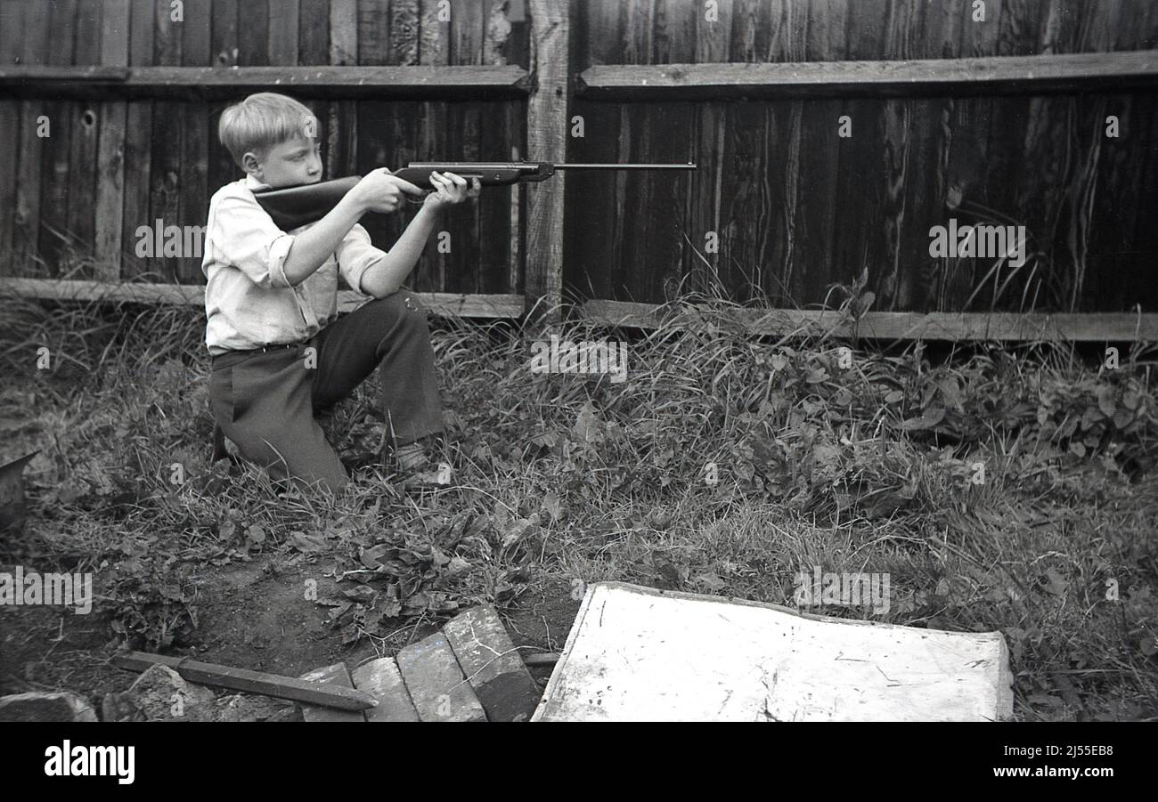 1960s, histórico, en el exterior en un jardín trasero, al lado de una valla de madera, un niño arrodillado, sosteniendo un rifle de aire en ambas manos en una posición de tiro o tiro, posiblemente un Diana modelo 27, Inglaterra, Reino Unido. Foto de stock