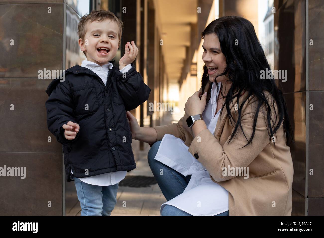 Mujer de pelo negro riendo y niño pequeño sonriente mirando la cámara en la calle cerca de la entrada. Niño sordo tratando de oír Foto de stock