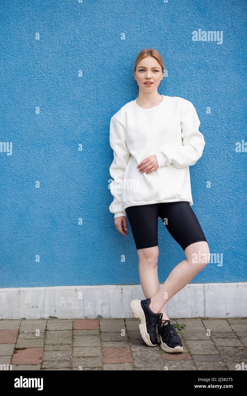 vista completa de mujer en sudadera blanca y pantalón corto de bicicleta  negro cerca de la pared azul al aire libre Fotografía de stock - Alamy