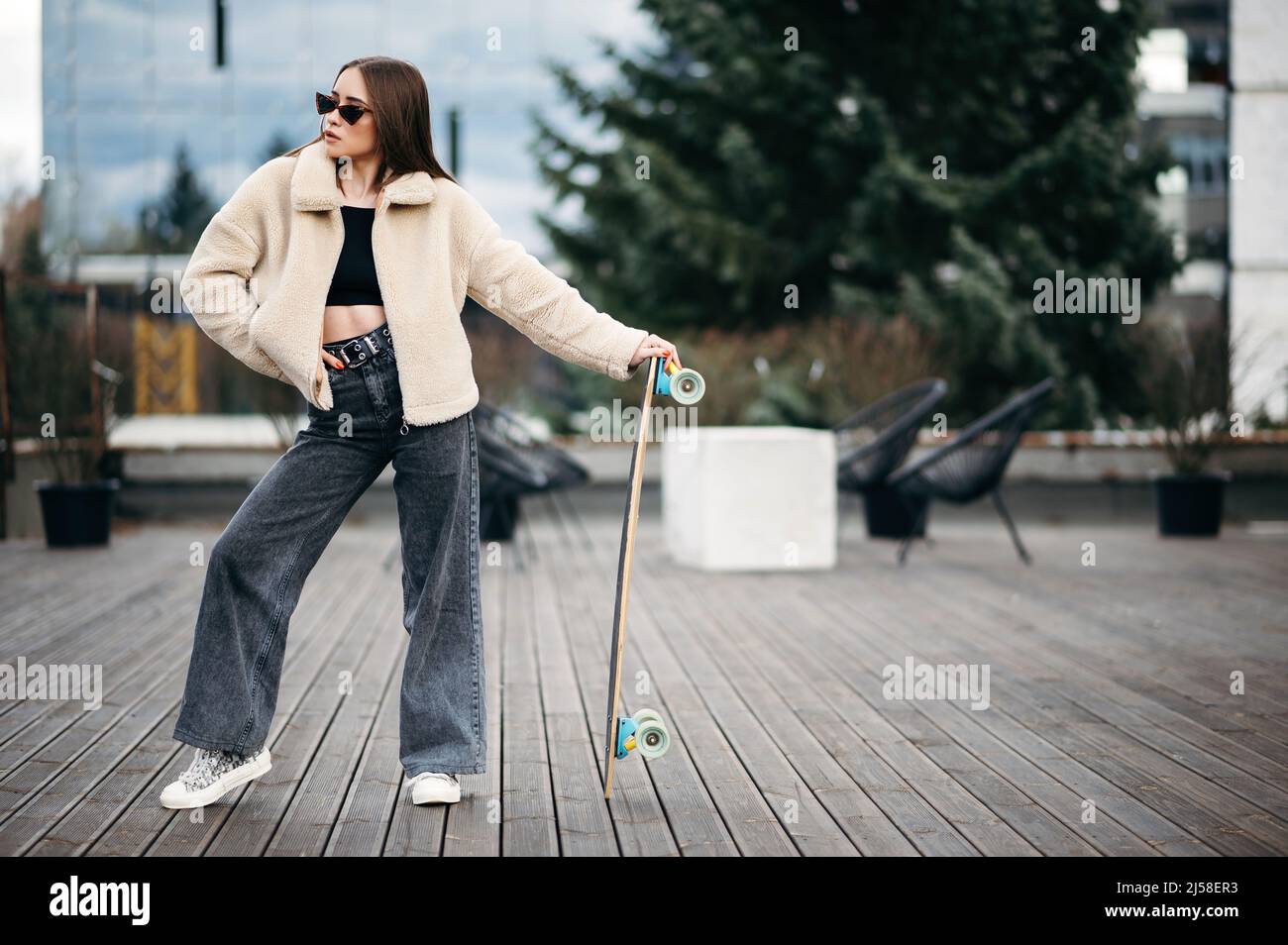 Atractiva mujer en ropa de calle de moda de pie con tabla de largo en la  azotea y mirando a un lado. Morena desenfadada con jeans oscuros, abrigo de  piel beige Fotografía