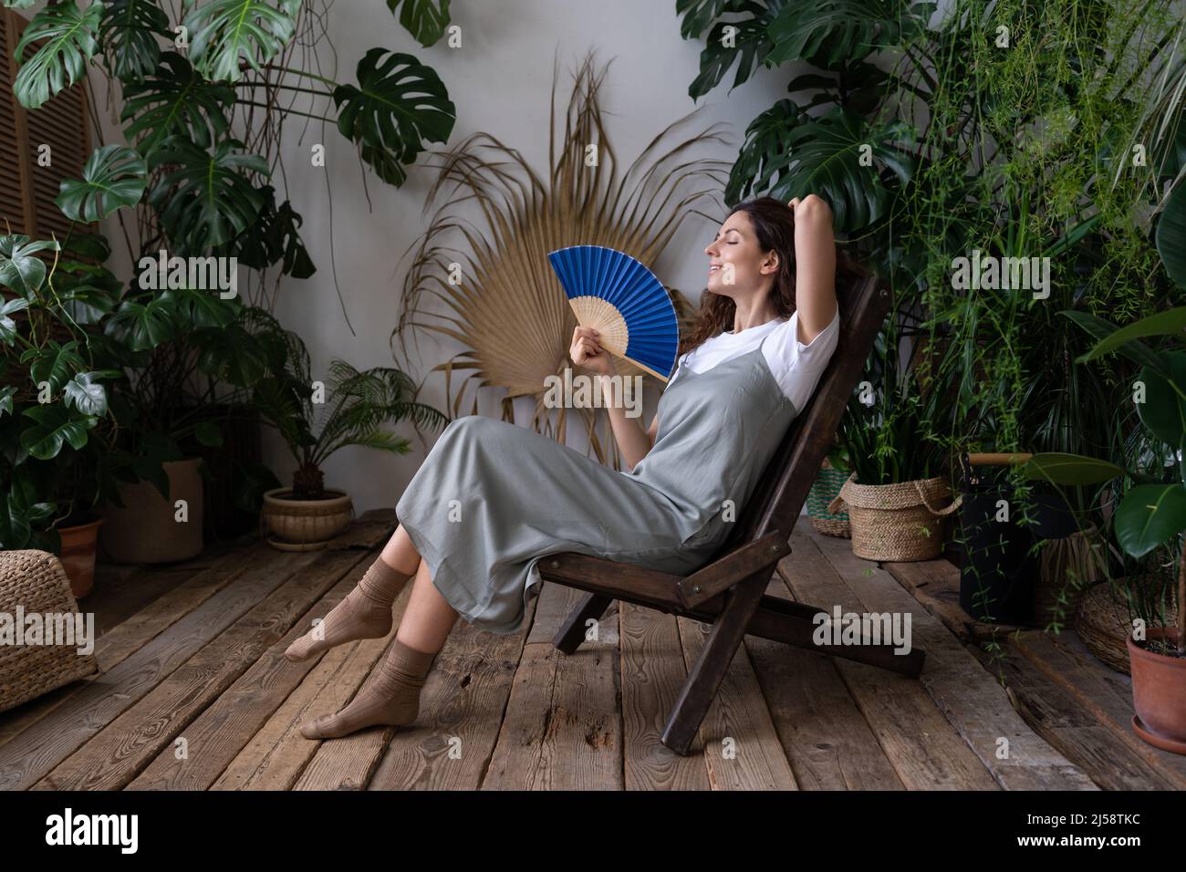 Joven mujer soñadora relajada utilizando un ventilador de papel mientras se relaja en el hermoso y refrescante jardín casero Foto de stock