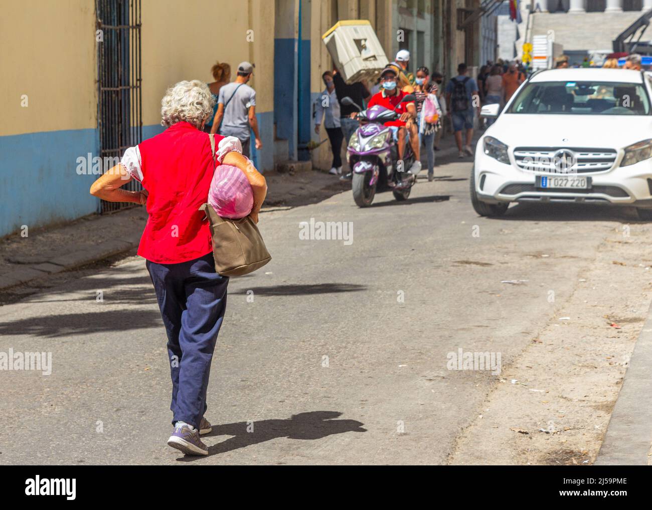 Una mujer cubana mayor con una blusa roja camina por una calle estrecha de la ciudad donde se ve a otras personas en su estilo de vida diario. Algunos llevan una caja Foto de stock