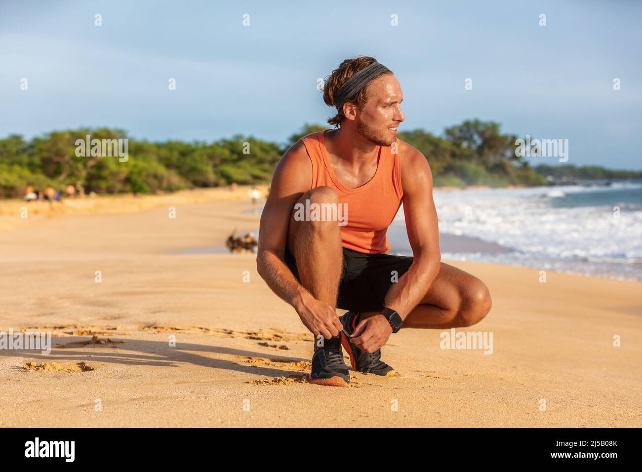 Hombre corredor de fitness SmartWatch atado zapatos para correr en la playa haciendo ejercicio de verano. Entrenamiento de running activo y saludable Foto de stock