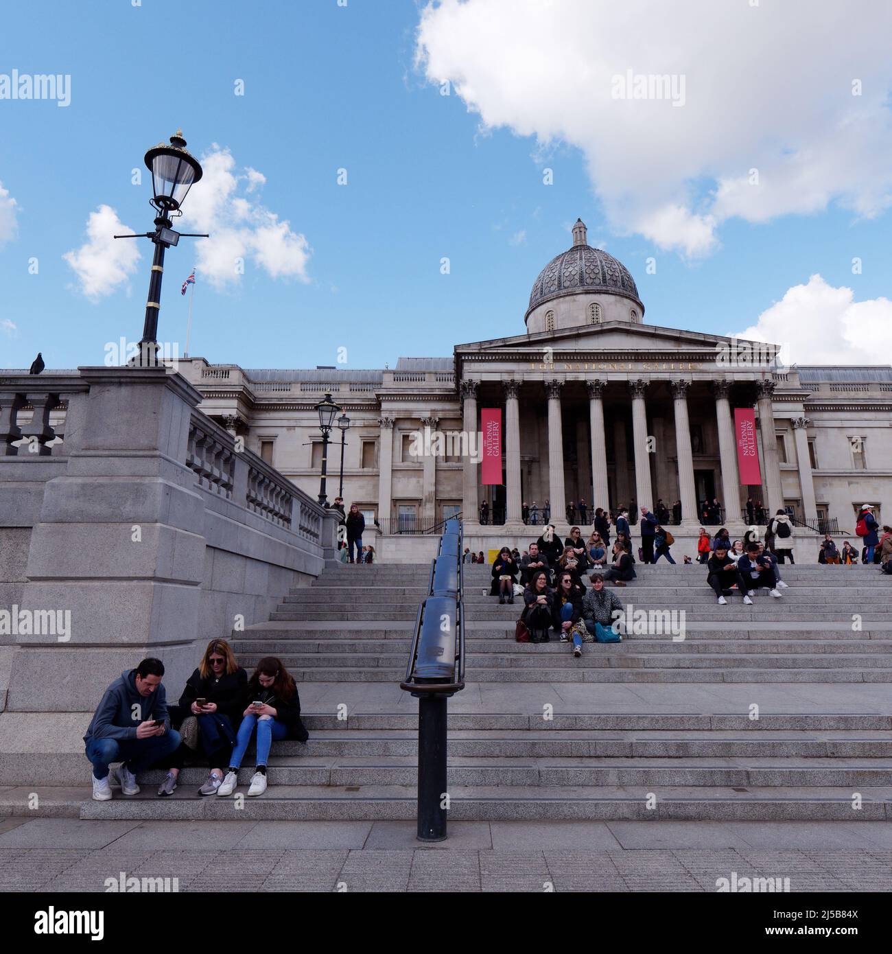 Los turistas se sientan relajándose y comprobando los teléfonos en las escaleras de Trafaglar Square en frente de la National Gallery de Londres. Foto de stock