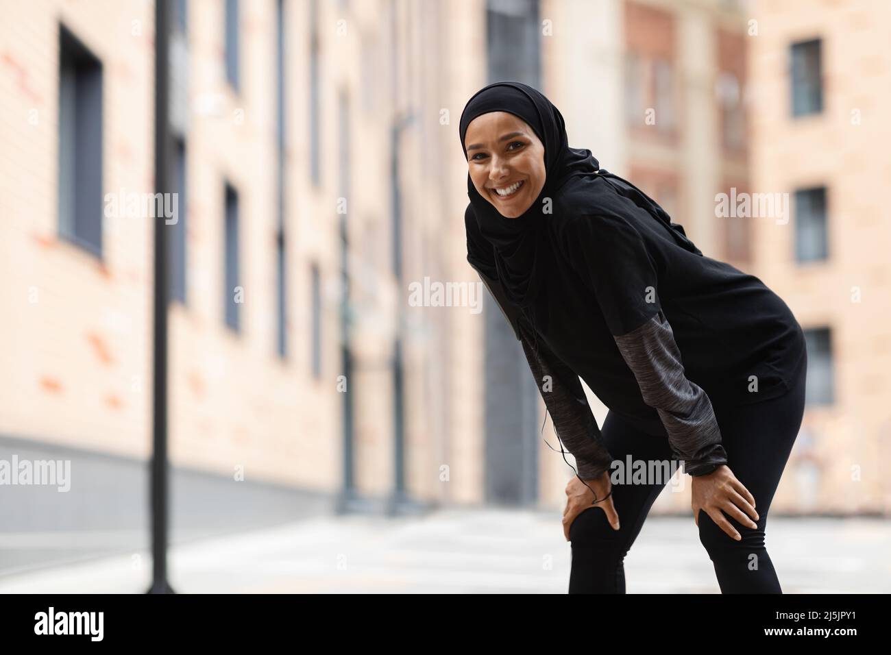 Alegre cansada joven musulmana atleta en ropa deportiva, hijab con smartphone está descansando después de correr Foto de stock