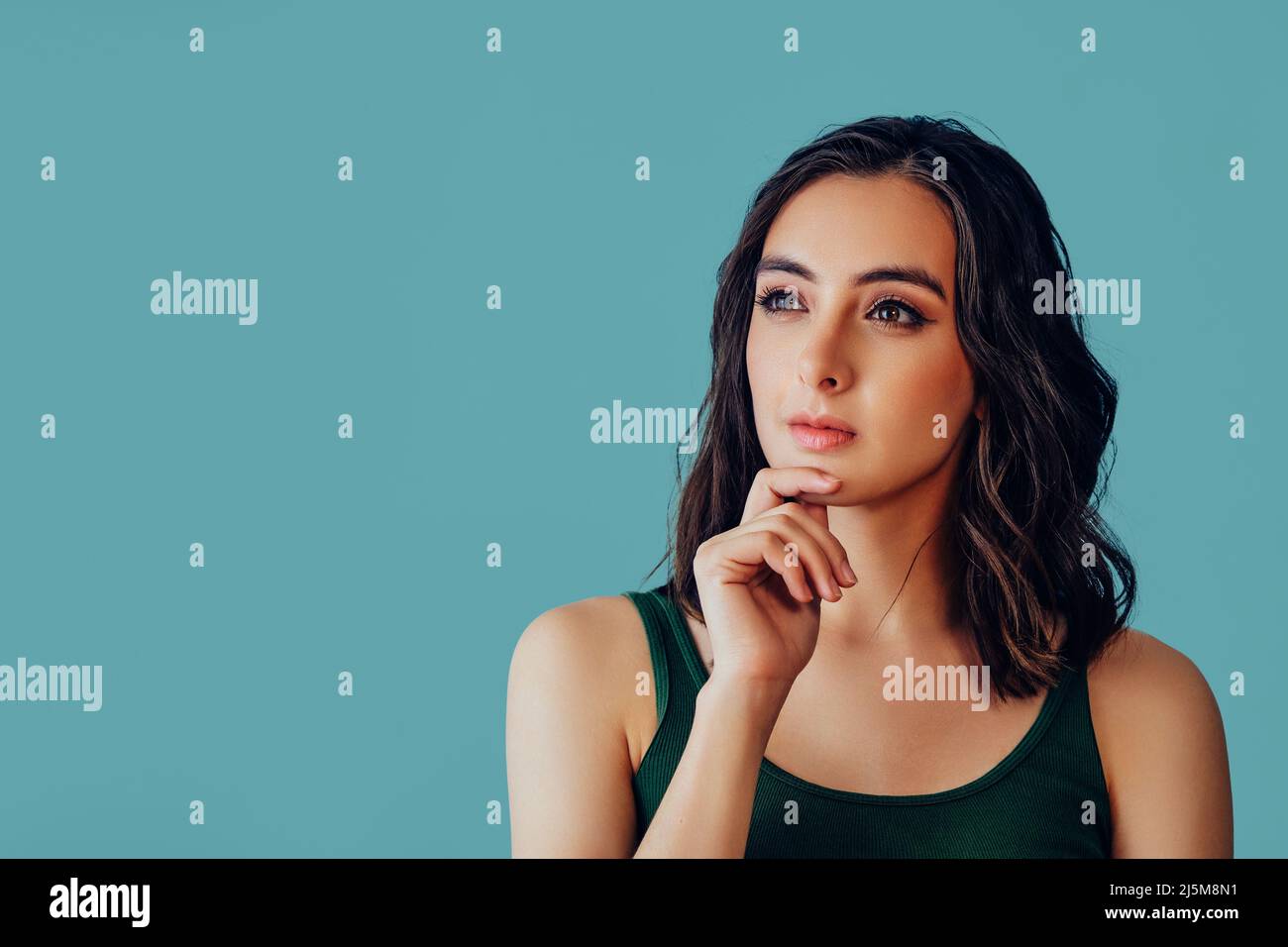 Mujer pensativa con la mano en la barbilla mirando para copiar espacio sobre fondo azul con verde tanque arriba retrato del estudio Foto de stock