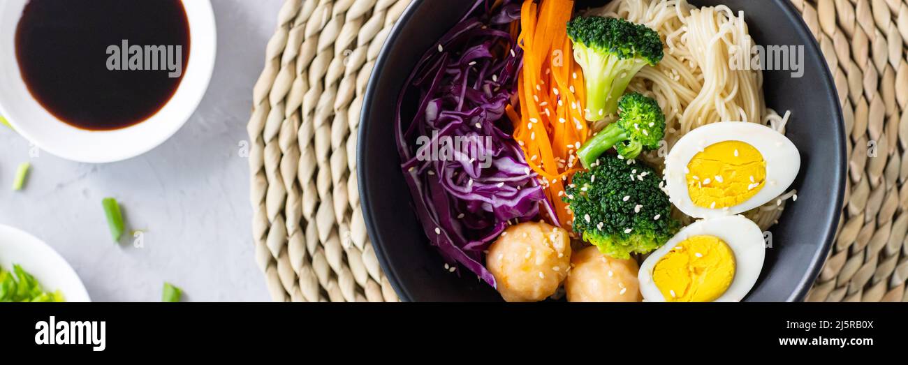 Alimentación y dieta saludables. Tazón negro de fideos asiáticos con salsa de soja, brócoli, col roja, zanahoria, bolas de camarón, Huevo y sésamo sobre hormigón gris Foto de stock