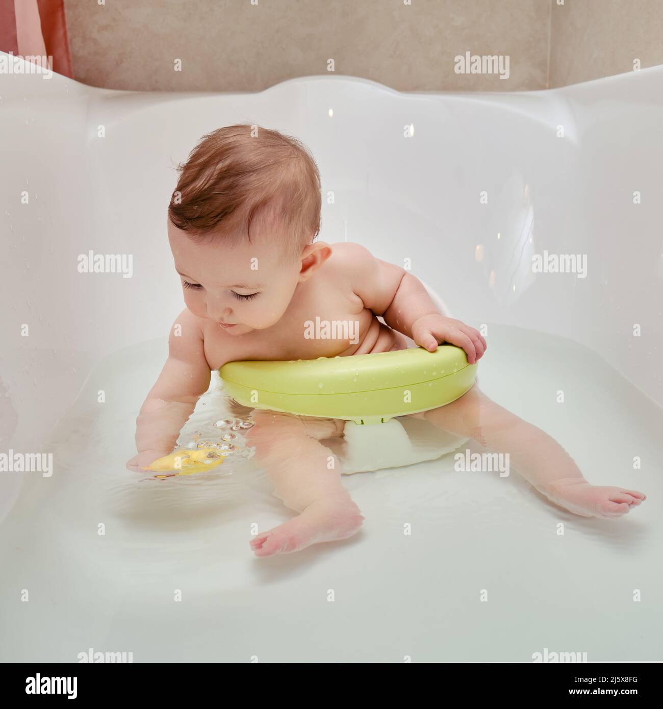 Un bebé feliz se baña en el mientras está sentado en una silla en una bañera blanca. Niño juega un juguete de pato amarillo mientras se baña, seis