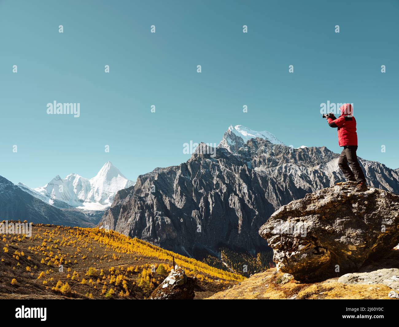 Hombre asiático de pie en la cima de la roca tomando una foto del pico de la montaña Yangmaiyong (o Jampayang en tibetano) en la distancia en Yading, condado de Daocheng, Sich Foto de stock