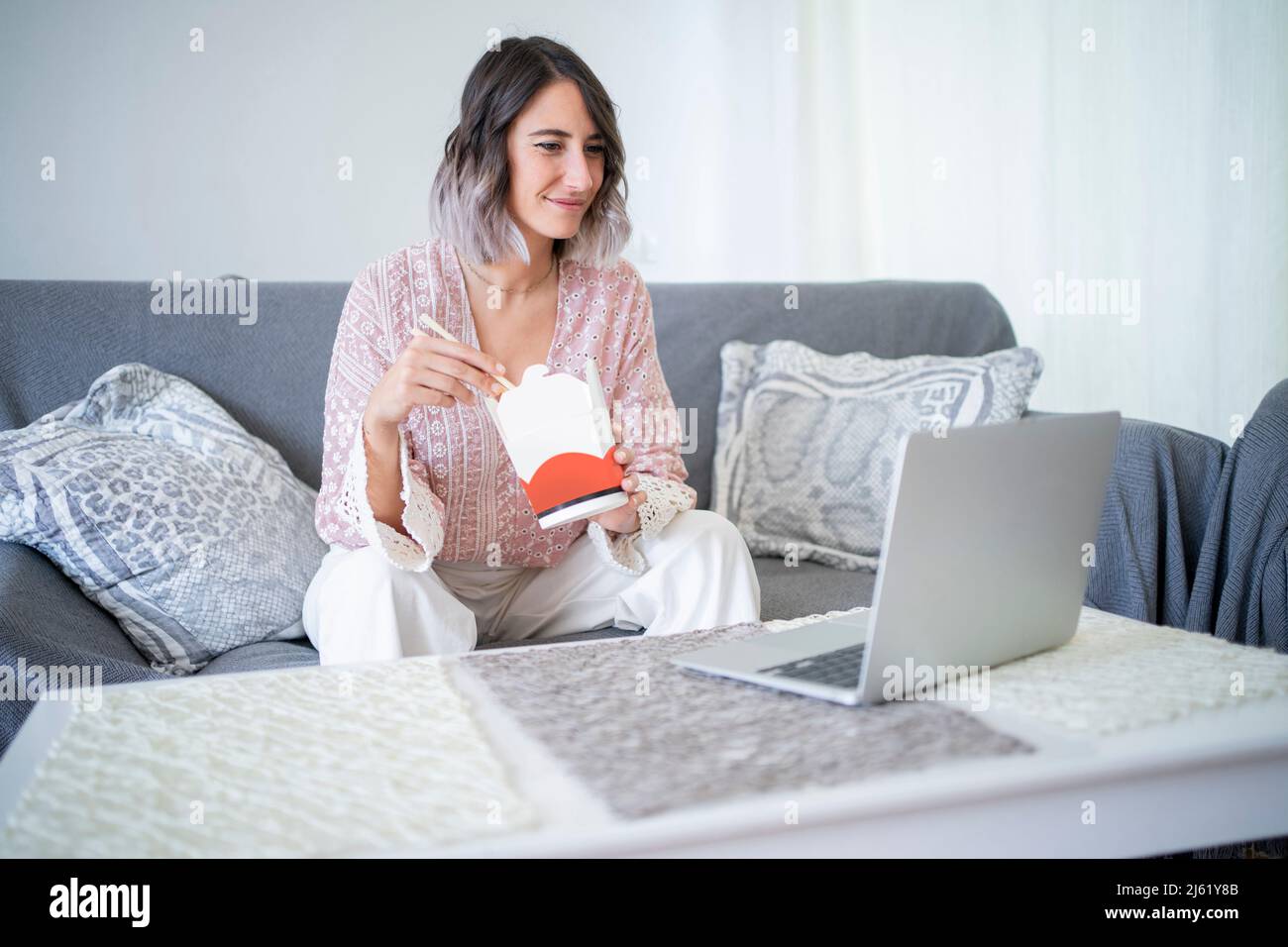 Mujer sonriente con comida para llevar viendo vídeo a través del portátil sentado en el sofá de la casa Foto de stock