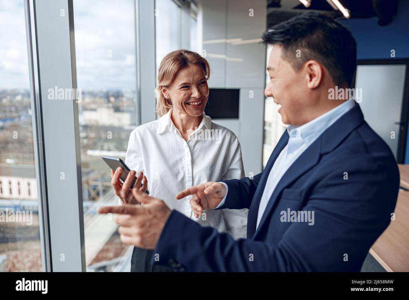 los empresarios asiáticos confiados hablan con su pareja madura mientras se encuentran cerca de la ventana panorámica Foto de stock