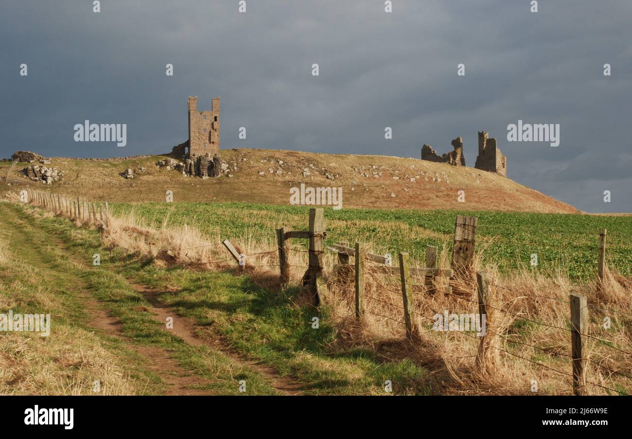 Imagen del paisaje que muestra cerca del lejano Castillo de Dunstanburgh desde el camino costero resaltado contra un cielo oscuro Foto de stock