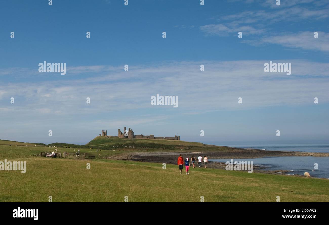Imagen del paisaje del lejano Castillo de Dunstanburgh en el umbral de Whin visto desde el camino costero de Craster con la gente caminando a lo largo del camino Foto de stock