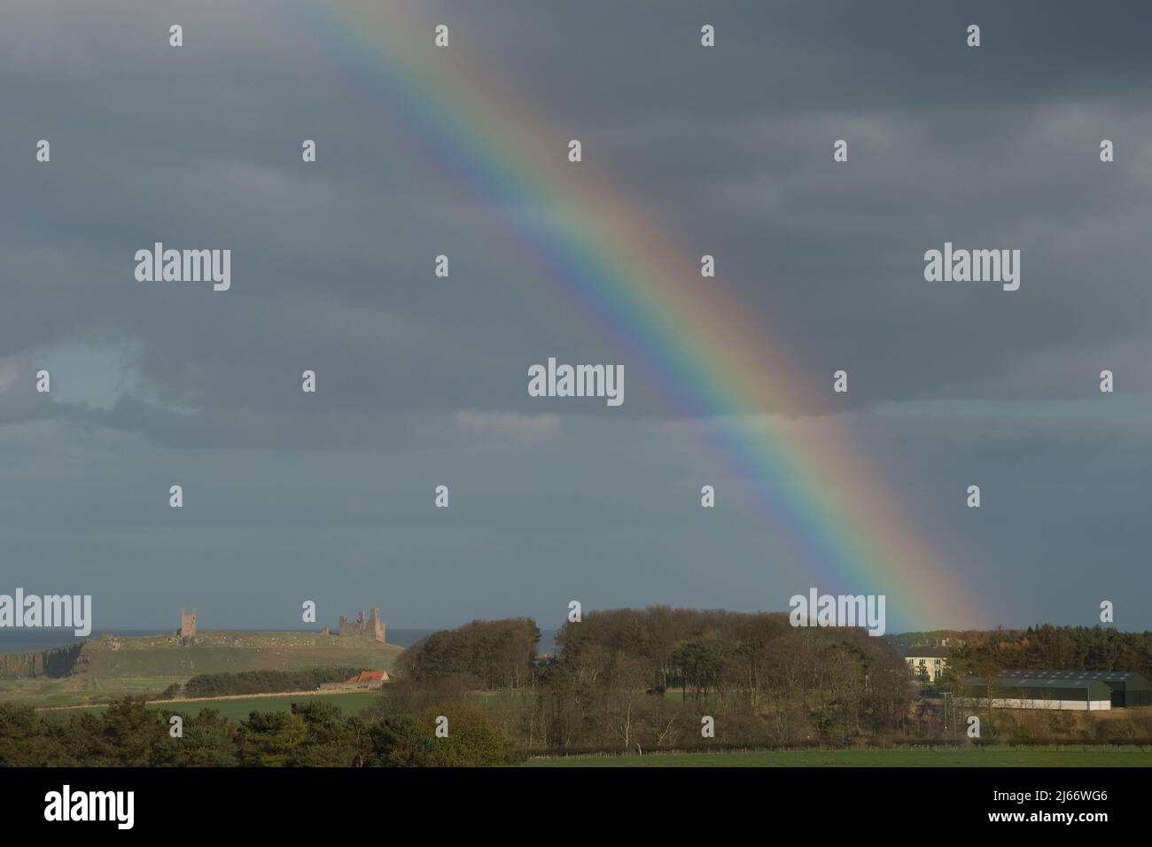 Paisaje a lo largo de la costa de Northumberland con la oruga de Dunstanburgh en la distancia cercana y un arco iris grande sobre el castillo a principios de la primavera Foto de stock