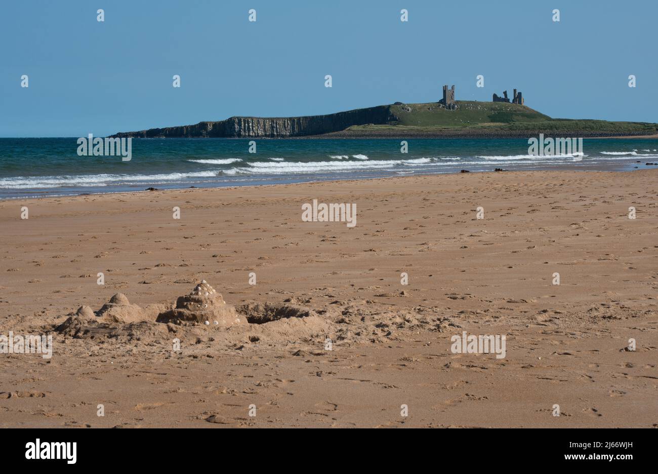 Un lejano Castillo de Dunstanburgh visto desde la playa de Embleton en un día de verano con un castillo de arena de un niño con foso actuando como un punto de vista en primer plano Foto de stock