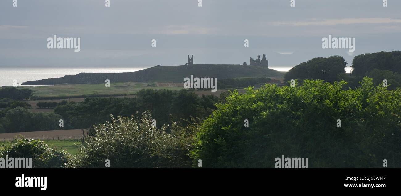 Un lejano castillo de Dunstanburgh visto a través de las tierras de labranza y contra el cielo con la luz del sol capturando e iluminando la superficie del mar detrás Foto de stock