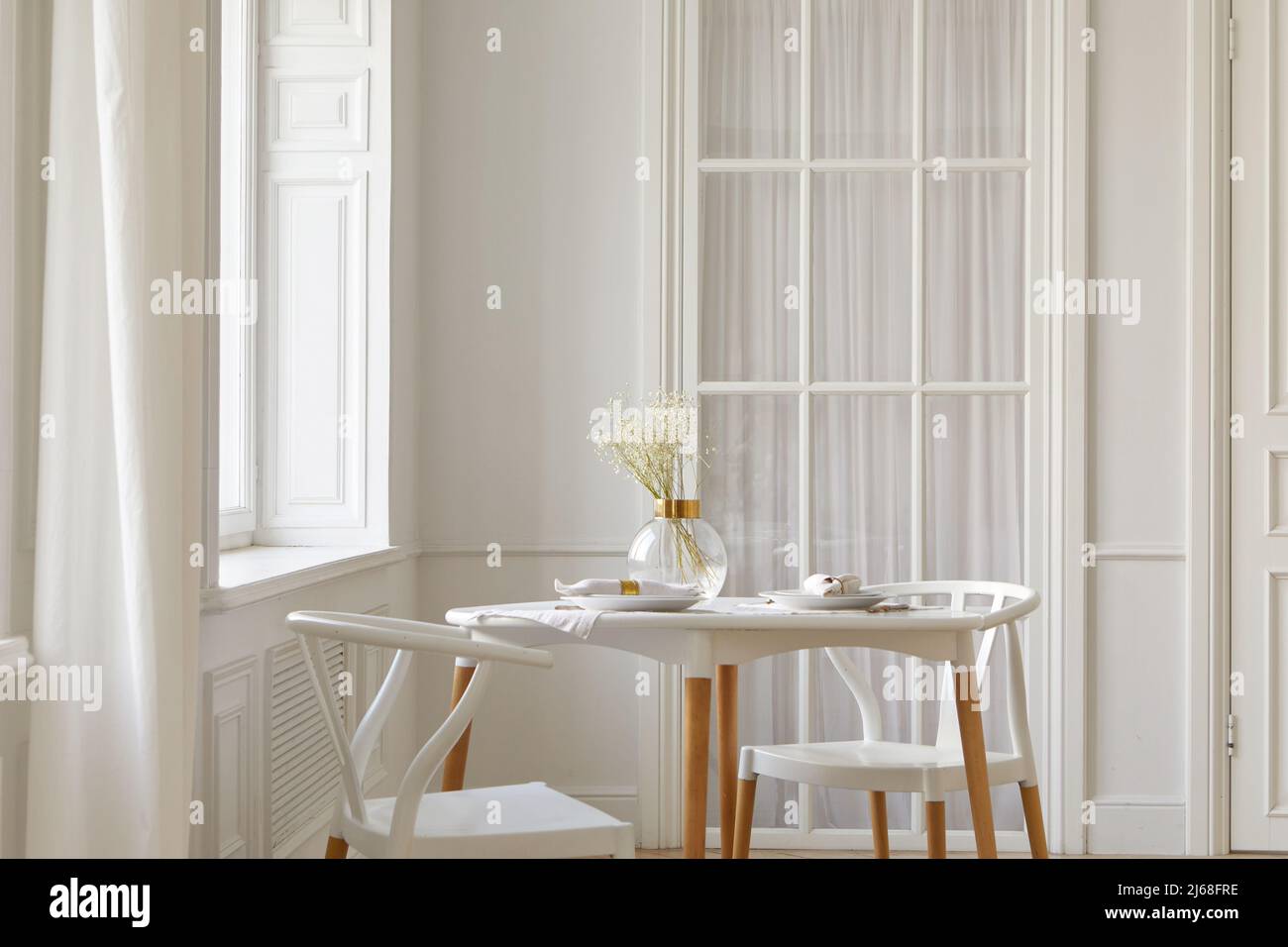 Jarrón de cristal con flores secas y vajilla colocada en la mesa cerca de sillas y ventana en una habitación blanca y espaciosa en casa Foto de stock