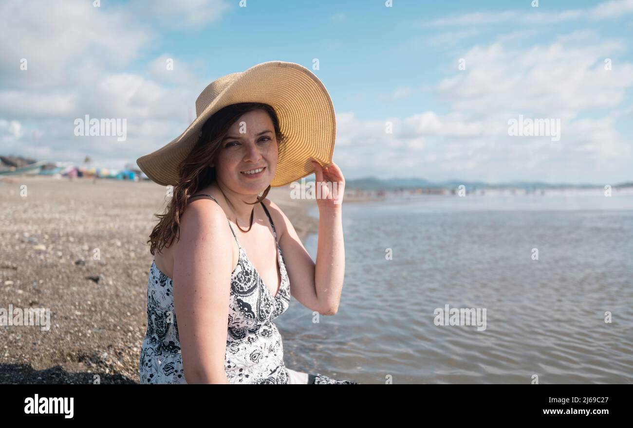Hermosa joven hispana sonriendo sentada sola en el borde de la playa mirando la cámara, sosteniendo su sombrero con su mano, usando un negro Foto de stock