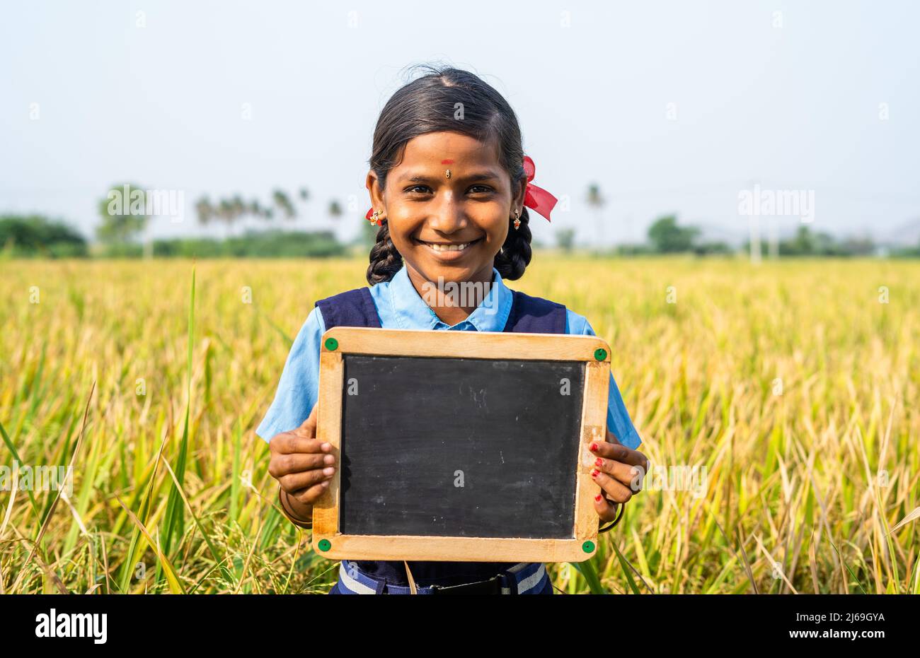 Feliz escuela niña sonriente sosteniendo pizarra vacía cerca del campo de arroz - concepto de educación, aprendizaje y empoderamiento de la infancia Foto de stock