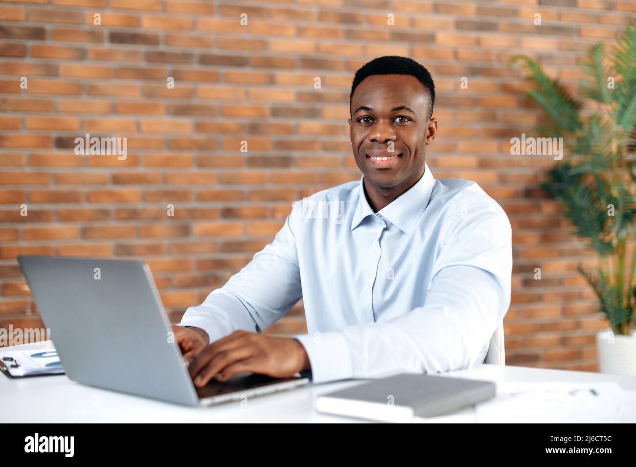 Retrato de un hombre afroamericano con éxito apuesto en camisa formal, hombre de negocios, director ejecutivo de una empresa, corredor, sentado en el lugar de trabajo en una oficina moderna, mirando la cámara, sonriendo amable Foto de stock