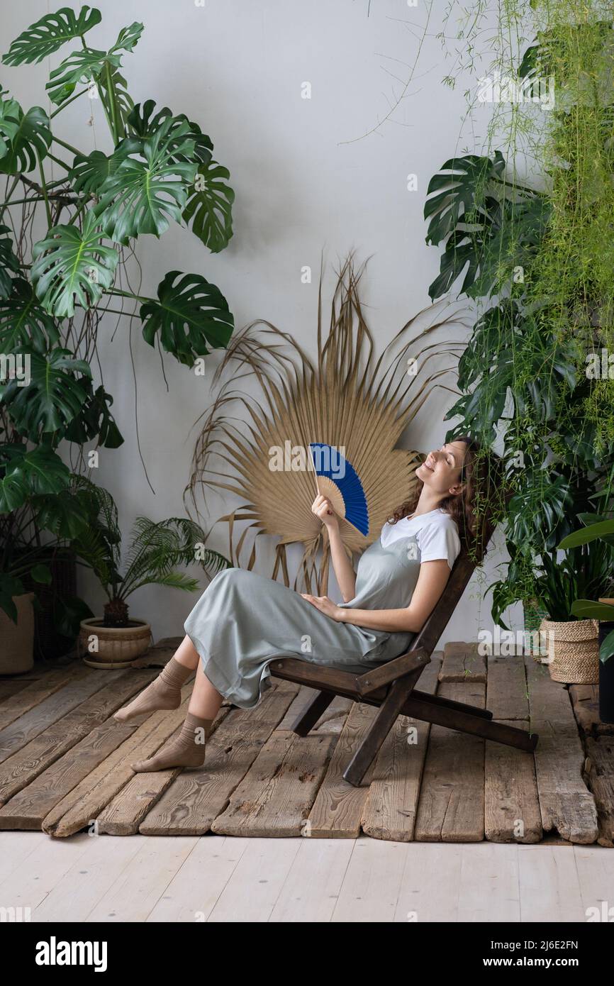Joven mujer soñadora relajada utilizando un ventilador de papel mientras se relaja en el hermoso y refrescante jardín casero Foto de stock
