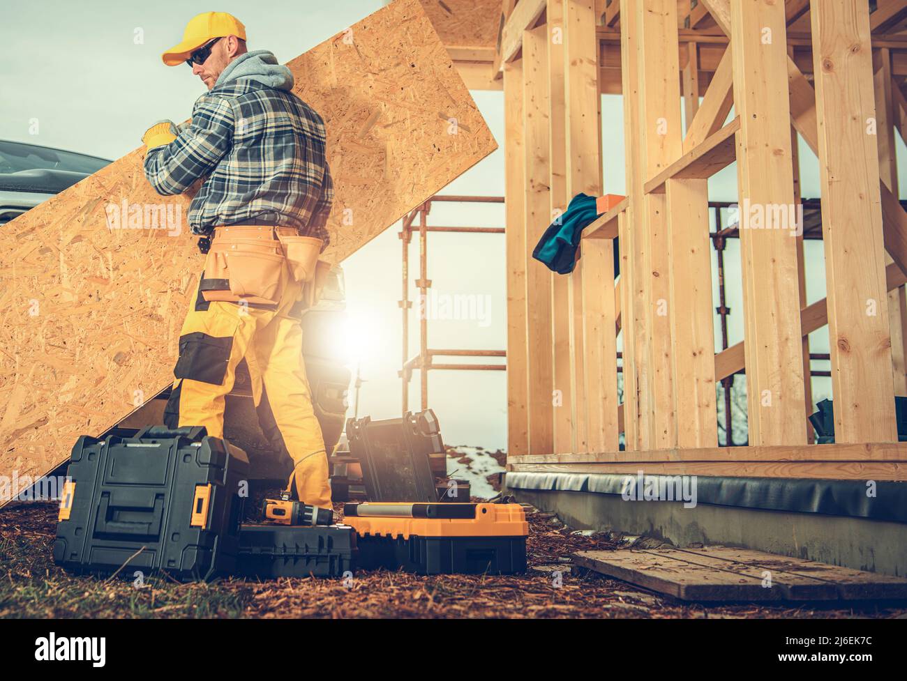 Trabajador de la industria de la construcción caucásica en sus 40s elementos móviles de contrachapado. Estructura de madera de la casa de esqueleto y herramientas de trabajo. Foto de stock