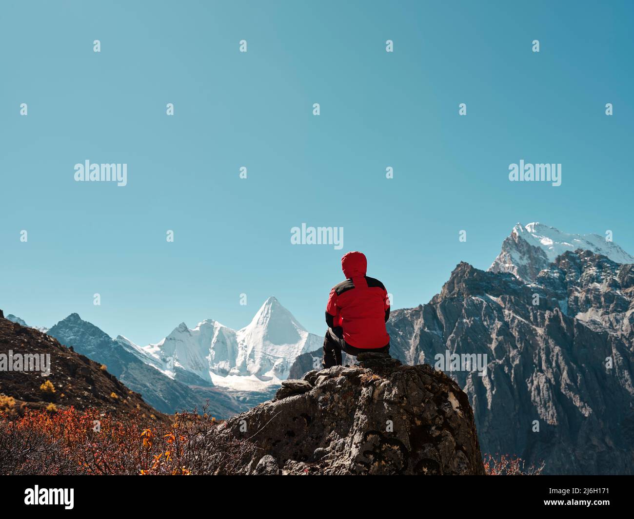 Vista trasera del hombre asiático sentado en la cima de la roca con Yangmaiyong (o Jampayang en tibetano) pico de montaña en la distancia en Yading, Daocheng County, Sic Foto de stock