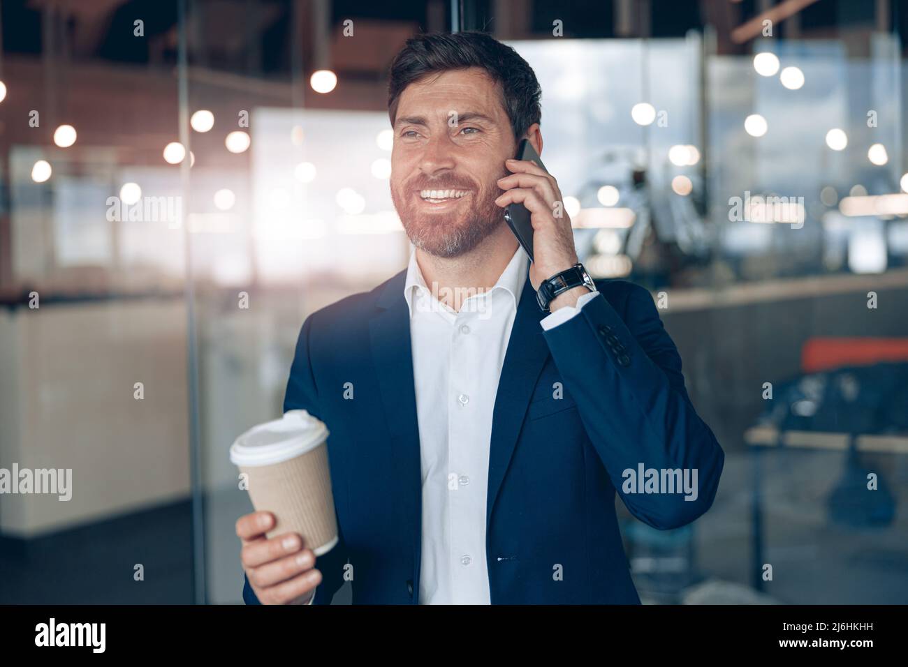 Un hombre de negocios guapo con una taza de café para llevar hablando por teléfono móvil con un socio de negocios Foto de stock