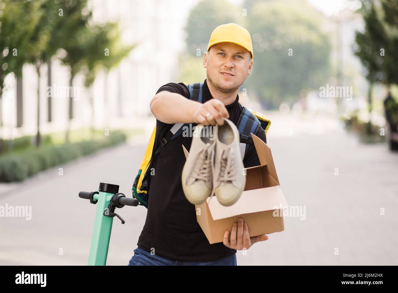 Hombre mensajero viaje en scooter electrónico con caja de cartón. Courier  abre la caja mirando a la cámara y saca sus sneakers. Concepto de entrega  de bienes de la tienda en línea