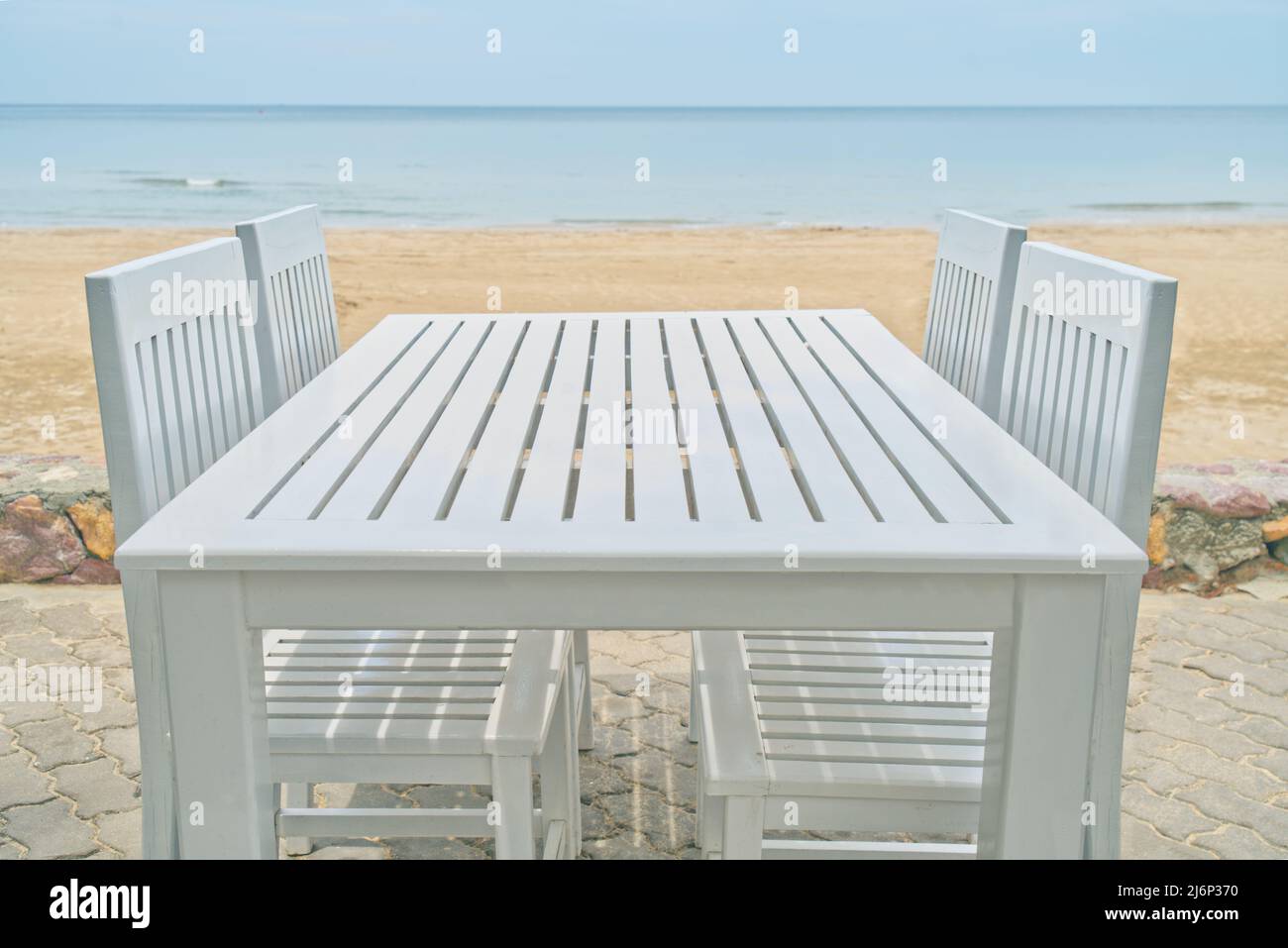 Hermosa mesa y sillas de madera en la playa, espacio vacío en la parte superior de la mesa, mesa de madera de color blanco, fondo borroso de la playa, el cielo y el océano. Foto de stock