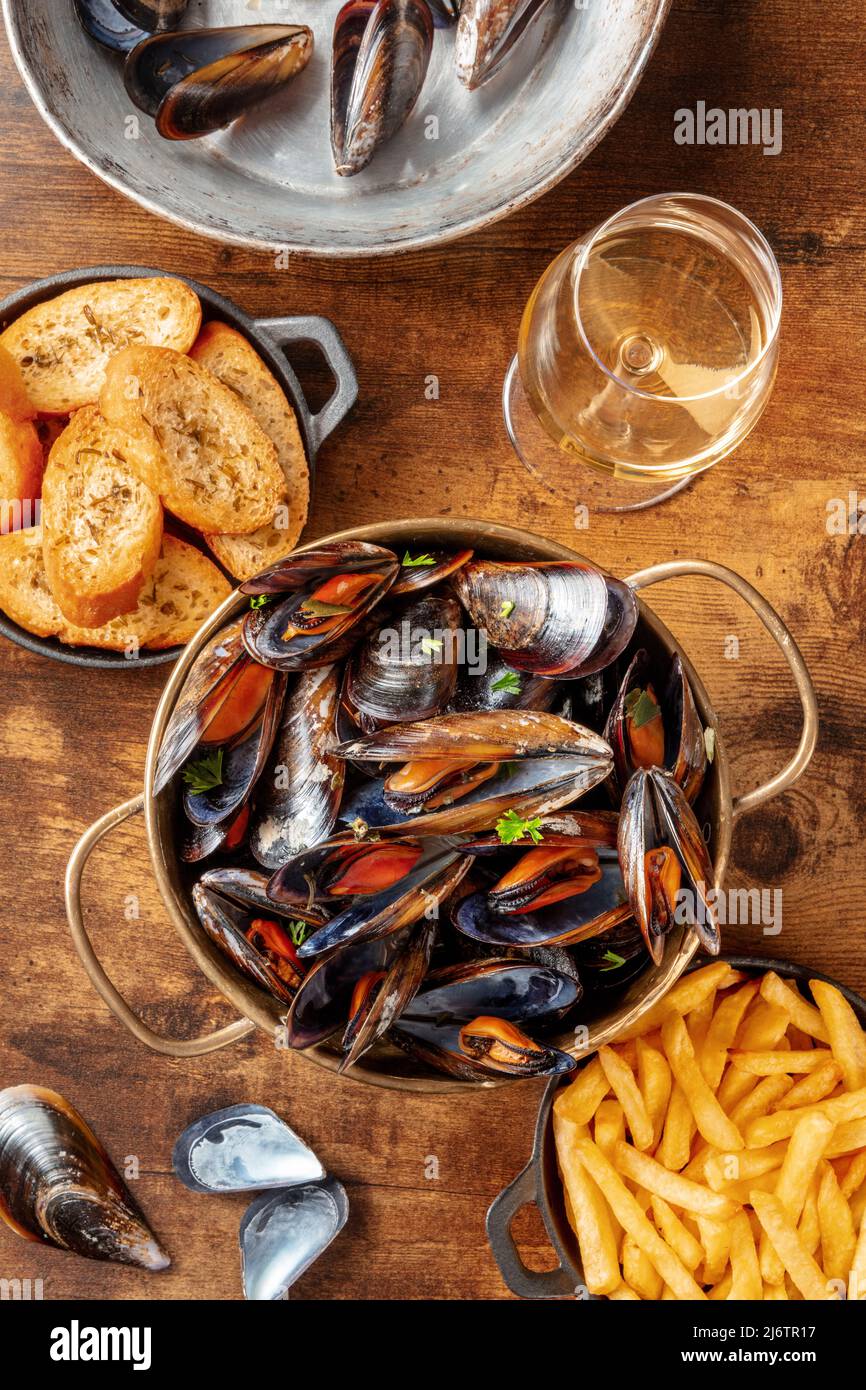 Mejillones belgas con patatas fritas y pan tostado, con vino blanco, sobre una mesa rústica de madera Foto de stock