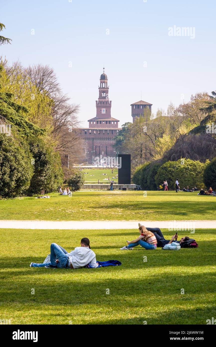 La gente disfruta del césped en el Parco Sempione (parque Simplon) en Milán, Italia, con vistas a las torres del Castello Sforzesco (Castillo Sforza). Foto de stock