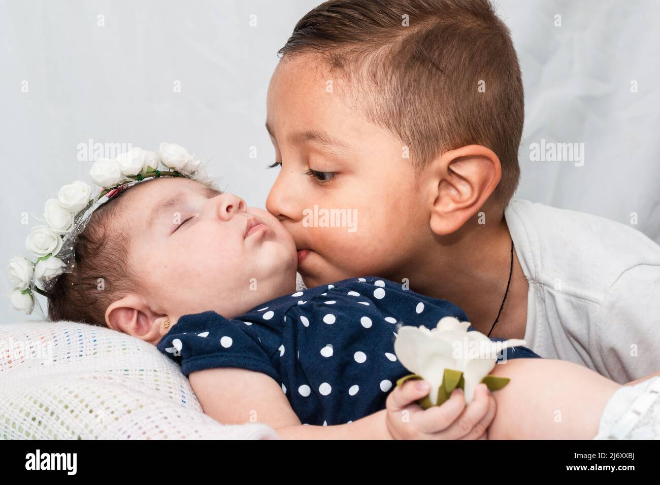 niño latino besando la mejilla de su hermana pequeña, el bebé está acostado sobre una almohada blanca, con los ojos cerrados, una corona de rosas blancas y sosteniendo una r Foto de stock