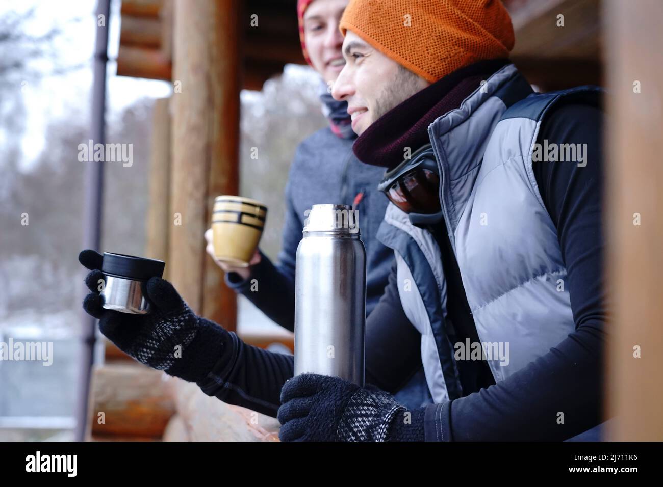 Amigos chatean después de esquiar. Se encuentran en el balcón de una casa de madera con ropa de invierno, beber té y relajarse. Portero de dos chicos en una estación de esquí en t Foto de stock