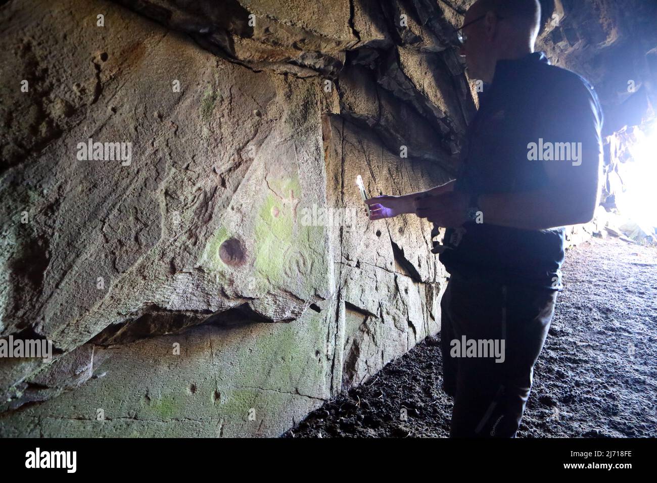 Hombre brillando una antorcha iluminando las primeras esculturas de roca cristiana y las posibles marcas de copa de bronce en Scoor Cave en la Isla de Mull, Escocia Foto de stock