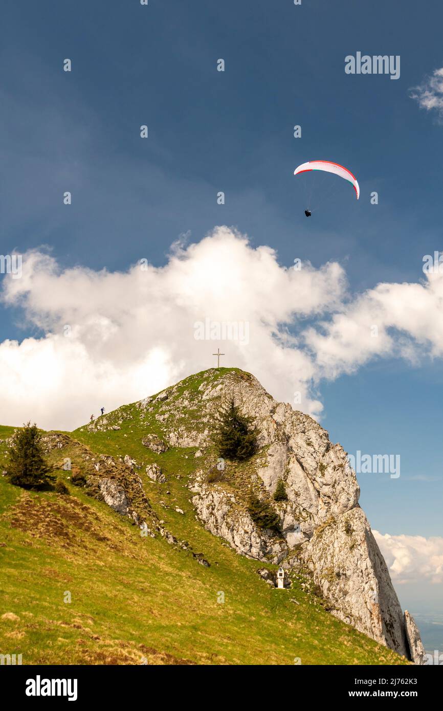 Un parapente rodea el Vorderer Kirchstein entre Brauneck y Benediktenwand en las estribaciones bávaras de los Alpes, el pequeño pico frente a una nube de luz en el cielo azul, mientras que el atleta vuela bastante cerca de él. Foto de stock
