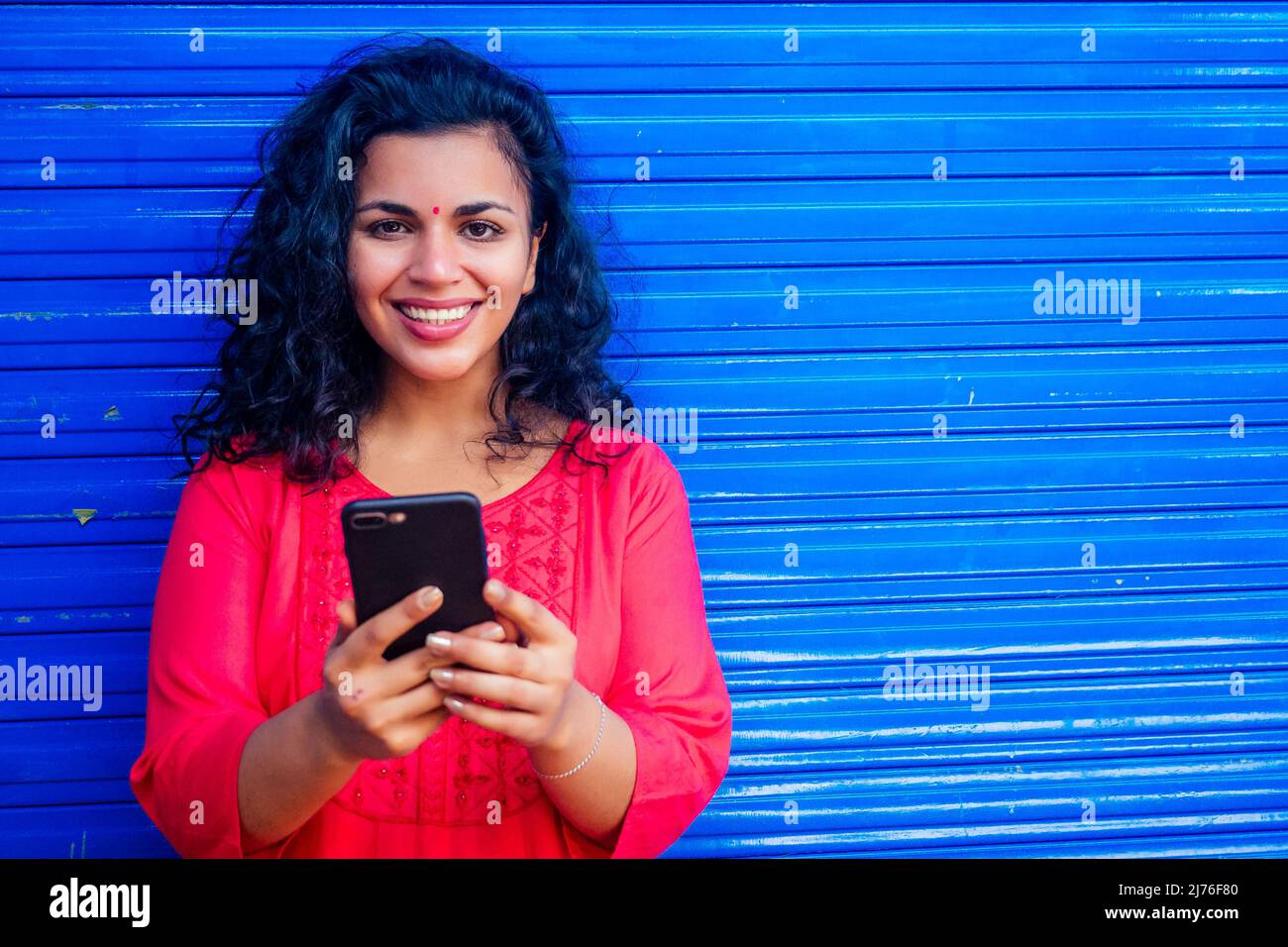 Atractiva hermosa feliz joven latina hispana mujer con bindi de teca en la frente sonriendo en la pared azul de fondo de calle sosteniendo smartphone moderno Foto de stock