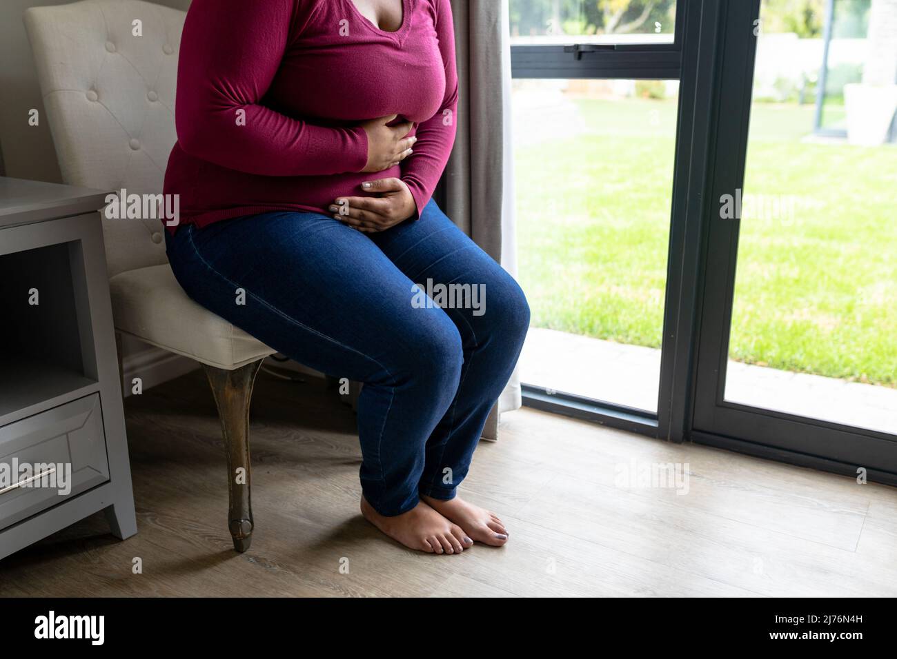 Sección baja de la mujer afroamericana de mediana edad que sufre de dolor de estómago mientras está sentada en la silla Foto de stock