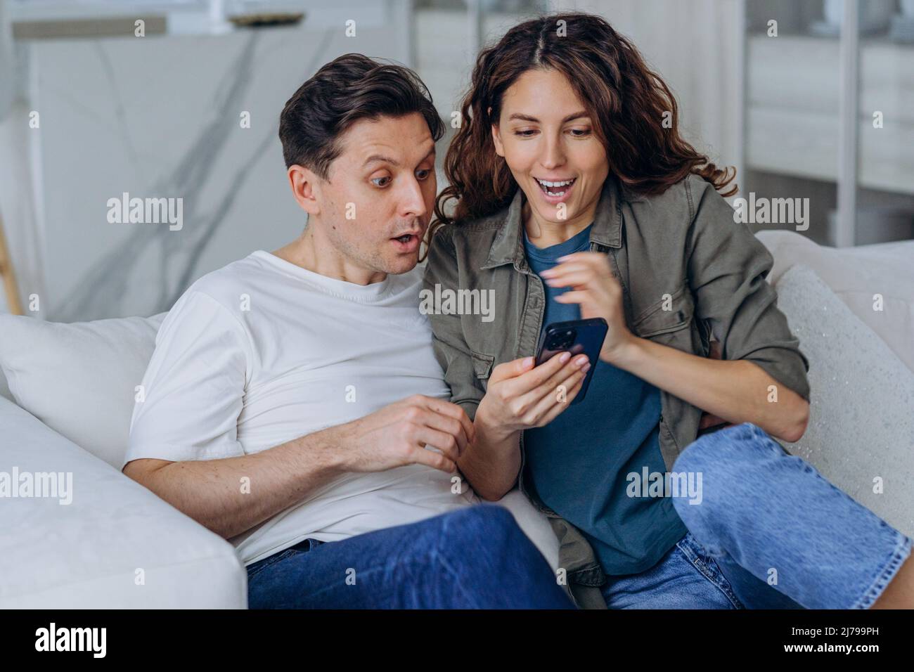 Emocionado mujer casada con pelo largo rizado y hombre morena sonrisa feliz leyendo buenas noticias en Internet a través de un smartphone sentado en el sofá Foto de stock