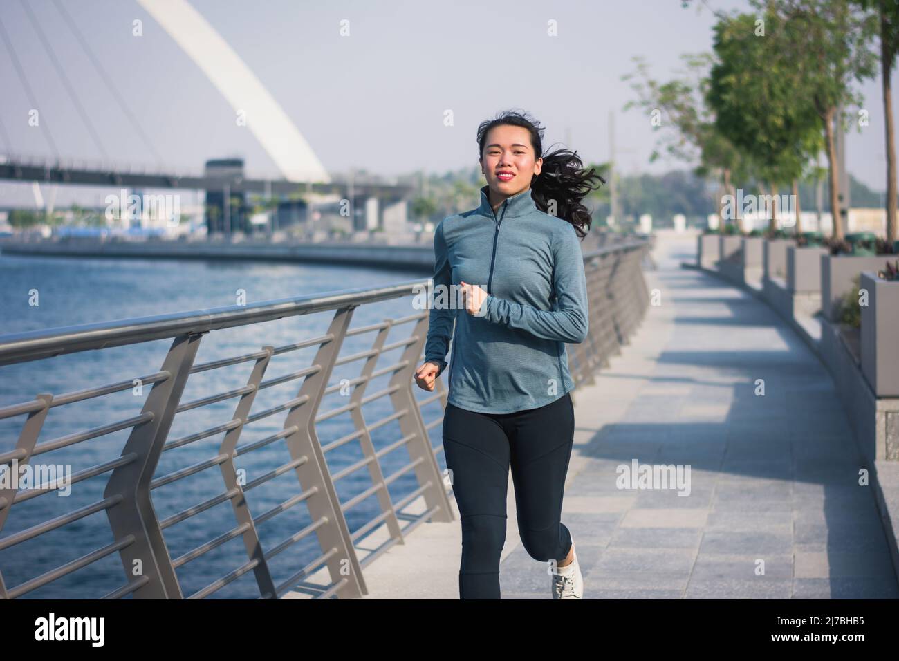 Forma mujer asiática corriendo en un ambiente moderno de la ciudad junto al agua en un paseo marítimo en Dubai. Jogging y estilo de vida saludable abstracto Foto de stock