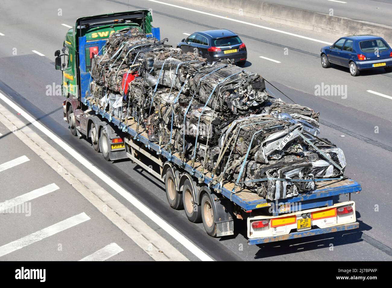 Vista trasera lateral aérea de carrocerías de chatarra triturada cargadas en un remolque articulado de plataforma plana remolcado por un camión hgv que circula por la carretera de la UK Foto de stock