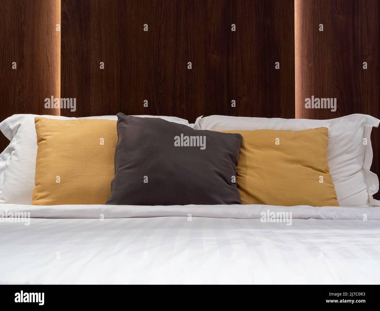 Conjunto de almohadas limpias, marrón, amarillo y blanco, manta y sábana en la habitación del hotel con cabecero de madera oscura y luz detrás. Ropa de cama en el Foto de stock