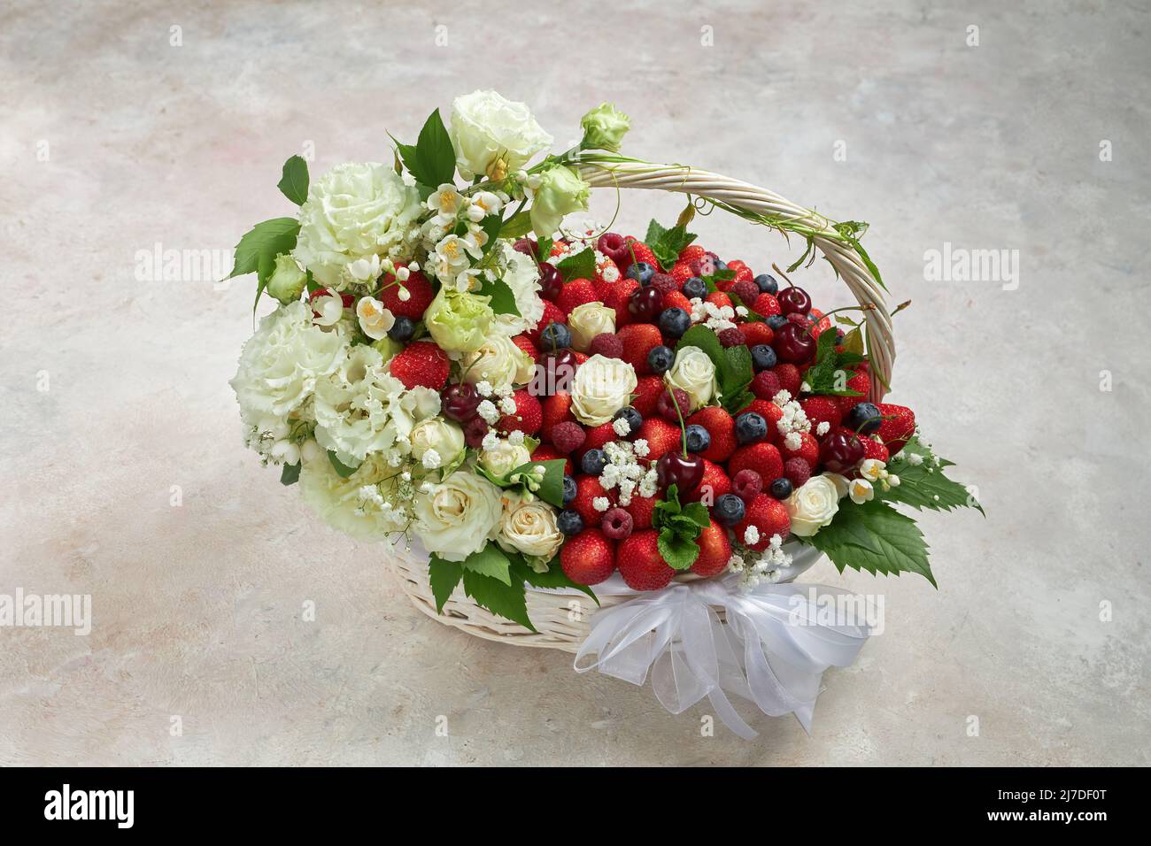 Cesta grande llena de fresas, frambuesas, arándanos, cerezas, decorada con flores blancas y hojas de menta Foto de stock
