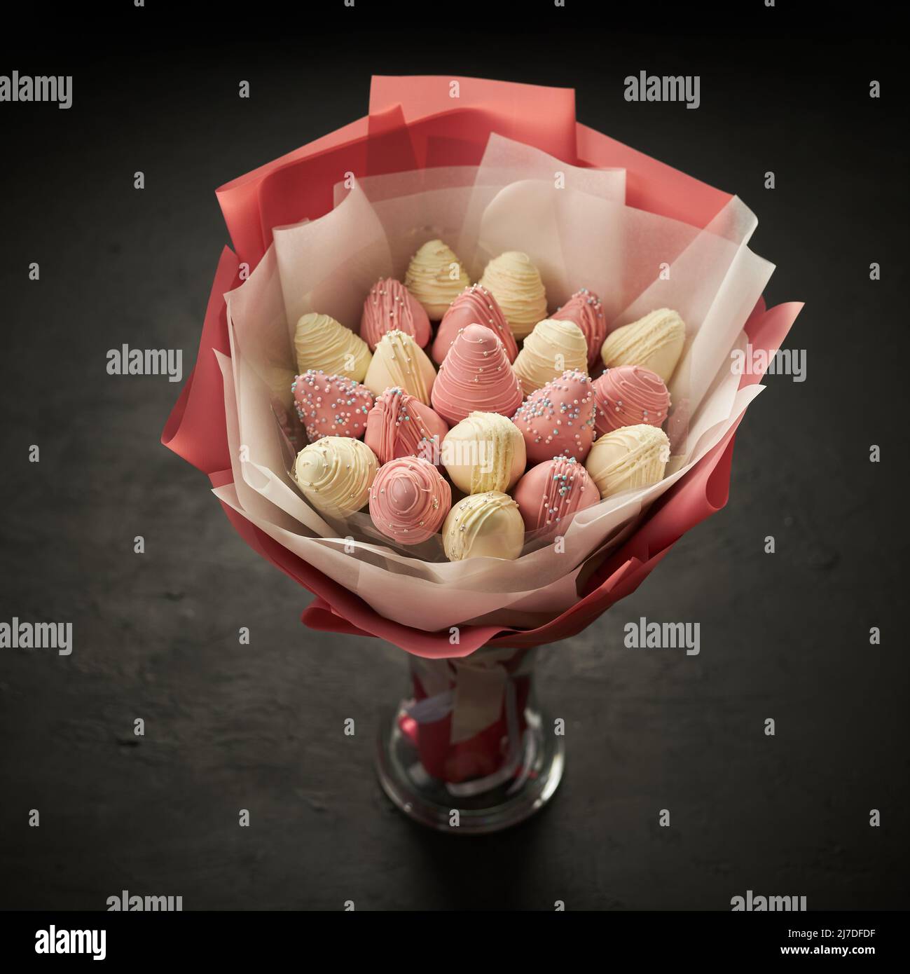 Un ramo de fresas maduras cubiertas de chocolate blanco y rosa se coloca en un jarrón sobre fondo negro Foto de stock