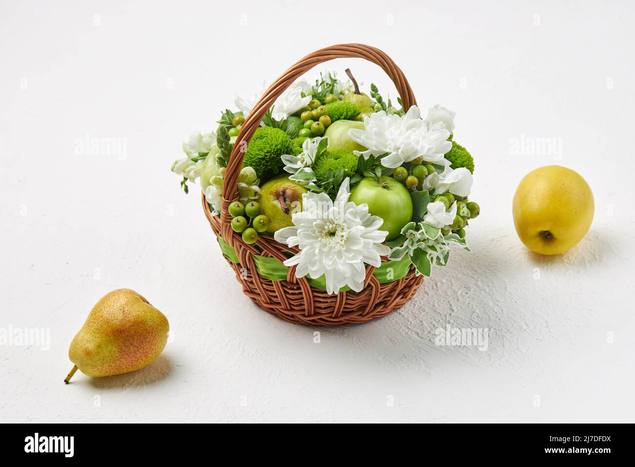Cesta de mimbre llena de frutas y flores blancas, así como una pera y una manzana sobre un fondo blanco Foto de stock