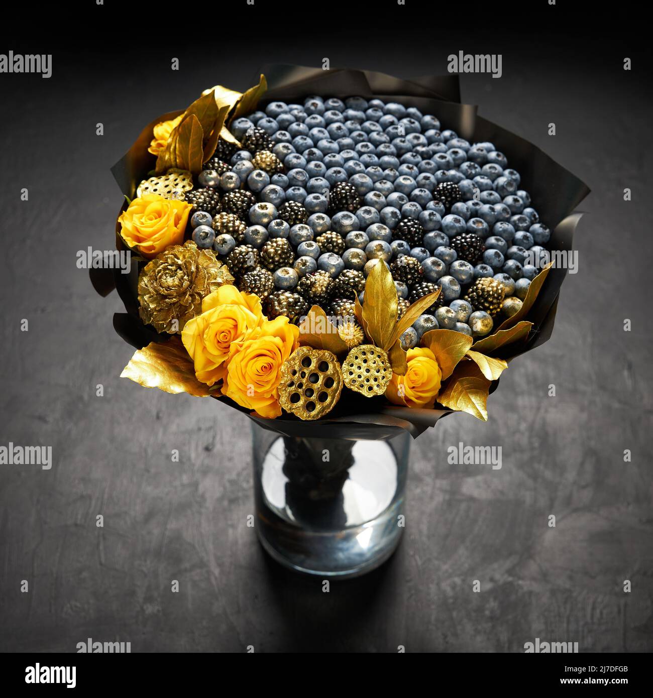 Fresco bouquet de arándanos y moras decoradas con rosas amarillas y hojas de oro de pie en un jarrón sobre fondo negro Foto de stock
