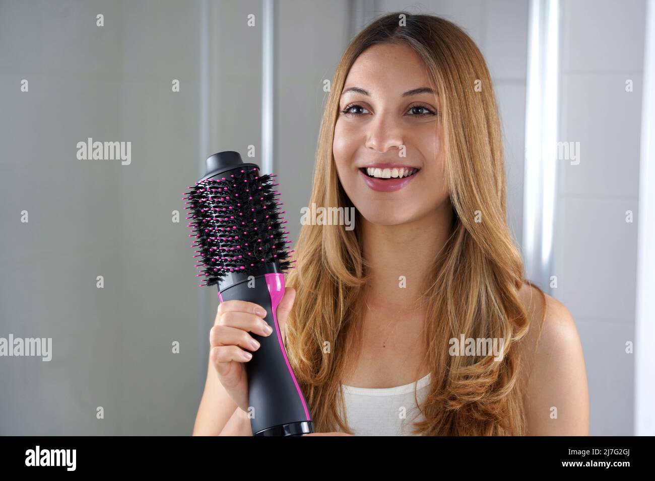 Hermosa niña sostiene el cepillo redondo secador de pelo para dar forma al  cabello en casa. Mujer joven que muestra salón de un paso secador de pelo y  volumizer Fotografía de stock -