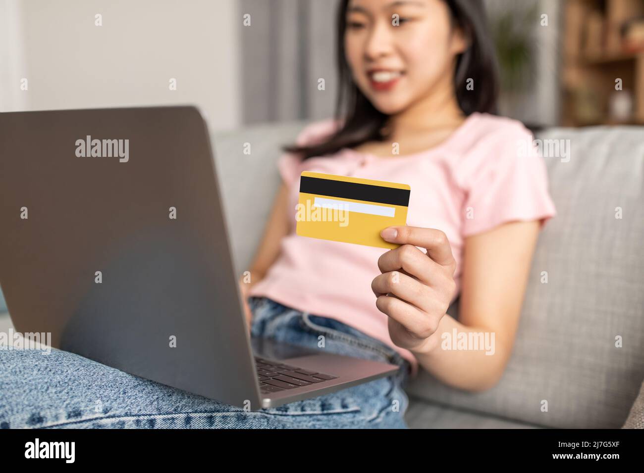 Hacer transacciones financieras. Joven dama asiática sosteniendo tarjeta de crédito, sentado en sofá con ordenador, enfoque selectivo Foto de stock